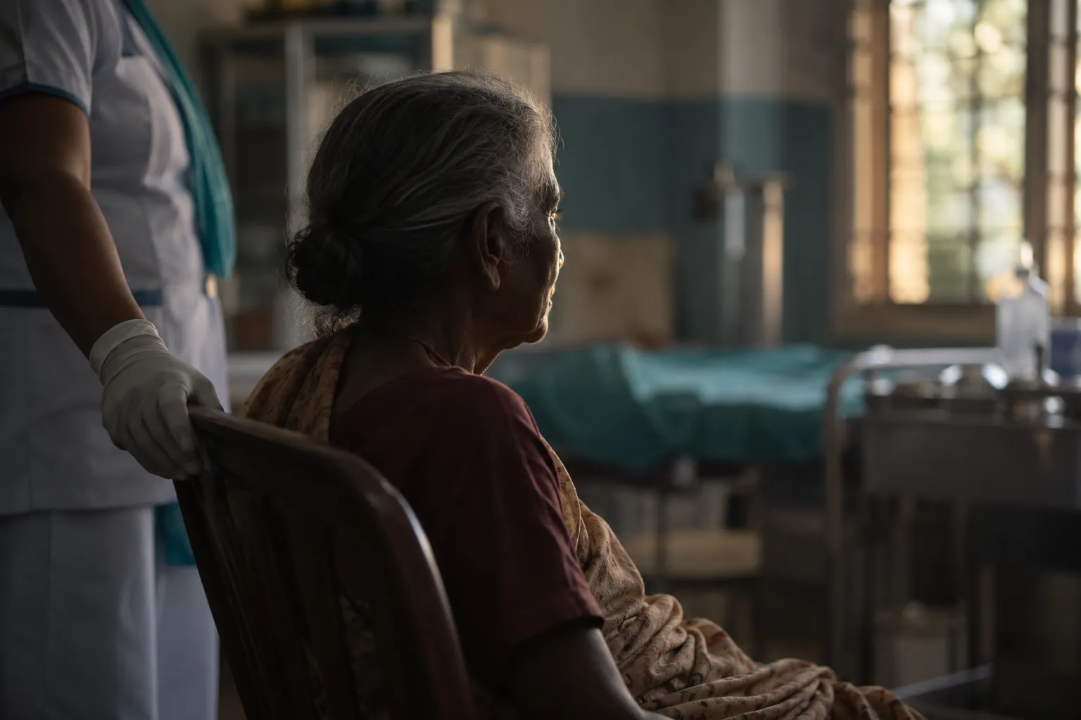 An elderly patient sits in a rural health centre, looking out of a window, with a nurse's gloved hand on the chair back.